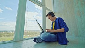 Happy boy in headphones with a laptop at home. School boy is sitting by the window and playing video games joyfully. Gaming concept.
