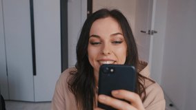 Young beautiful brunette woman using smartphone and drinking coffee at home. Smiling millennial girl looking at mobile phone screen, typing message in social network, shopping online, web surfing.