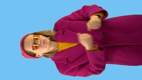 Beautiful young woman in a red hat and coat, in fashionable glasses, dancing on a blue background. Fashionable look, style, video for advertising. Vertical shot.