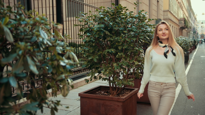 Young blonde woman walks through the ancient Italian city of Milan, looking at architecture, dressed in blue sweater, gray jeans and scarf, next to small trees in vases