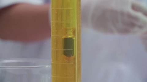 Hydrometer floats in the cylinder test jar filled with yellow chemical substance to measure density of liquids (specific gravity test) while a scientist pours substance from a beaker to a test tube.