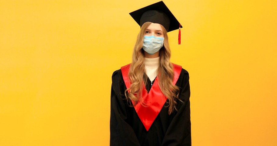beautiful female graduate, wearing a medical protective mask, with a diploma on an isolated yellow background. Graduation, quarantine, coronavirus Royalty-Free Stock Footage #1051409686
