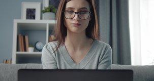 Close up of serious young woman in grey sweater and eyeglasses working on laptop while sitting on couch. Concept of remote work and digital device.