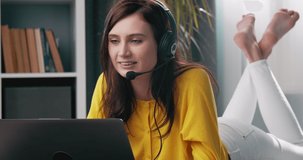 Smiling beautiful girl in headset chatting with friends during video call on laptop. Female with dark hair relaxing on floor and using modern devices.