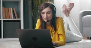Happy woman with brown hair relaxing on floor during video chat on laptop. Young lady in casual clothing using headset for online conversation at home.