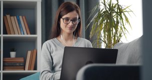 Pleasant happy female freelancer in eyeglasses working on laptop while sitting on couch. Beautiful woman in domestic clothing typing on computer at home