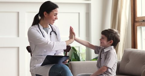 Smiling young female family doctor in uniform consulting little kid patient at checkup meeting at home. Friendly pediatrician giving high five to small child boy, celebrating good treatment results.