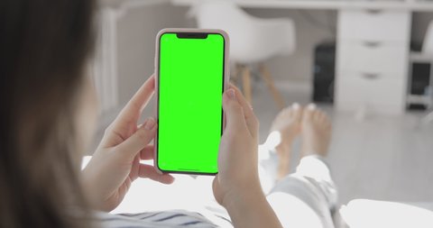 Back view over shoulder of young asian girl using smartphone with green screen laying down on bed while chatting with friend. iPhone 11 advertising mock-up. 2020 - Hong Kong, China