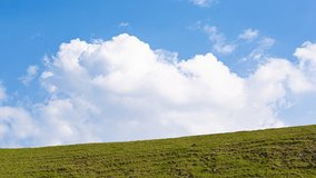 Fluffy white clouds flowing in the blue sky above a green hill on a warm day timelapse video. Bright vibrant colours, beautiful background. 