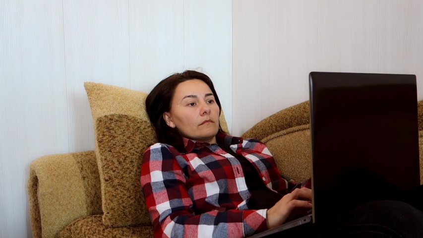A young woman in a plaid shirt lies on a sofa with a laptop.