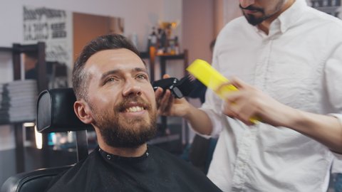 Young male barber shaving clients beard with electric trimmer. Handsome bearded man getting beard trimmed by hairdresser at barber shop. Confident man visiting hairstylist in salon