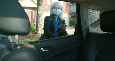 A mature woman as a ride sharing customer wearing a mask due to COVID19 checks the service app on her mobile phone as she gets into the vehicle.