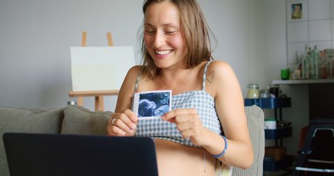 Happy young pregnant woman showing photo of her baby during online call with family by laptop computer while sitting on sofa in living room at home. Self isolation quarantine.