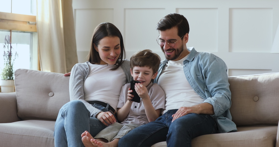 Happy little kid boy sitting on comfortable sofa between smiling parents, playing mobile game indoors. Smiling young family couple controlling child technology usage, relaxing together on couch. | Shutterstock HD Video #1051432597