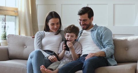 Happy little kid boy sitting on comfortable sofa between smiling parents, playing mobile game indoors. Smiling young family couple controlling child technology usage, relaxing together on couch.