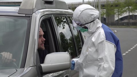 Health care worker in protective hazmat suit (and gloves, mask, and glasses) takes sample from coronavirus Covid-19 patient, through window of an SUV in a drive thru test clinic
