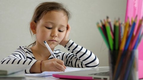 Little child girl is focusing on laptop screen and making notes in notebook during online lesson at home