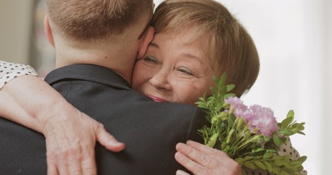 Close up view of smiling mature mother hugging her adult son while holding flowers in her hand. Man visiting his elderly mother at home. Concept of family, mothers day and birthday