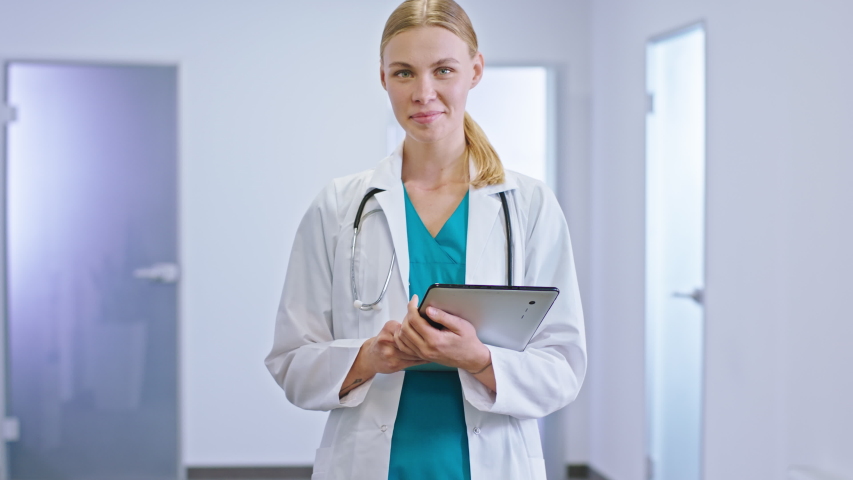 Charismatic lady doctor in the modern hospital corridor holding her personal digital tablet she analyzing the diagnostic of the patient and loo straight to the camera. Shot on ARRI Alexa Mini