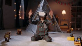 Cute little boy wearing pajamas is diving into virtual reality world before bedtime, using vr 3d headset, playing video games - modern technology concept 4k footage
