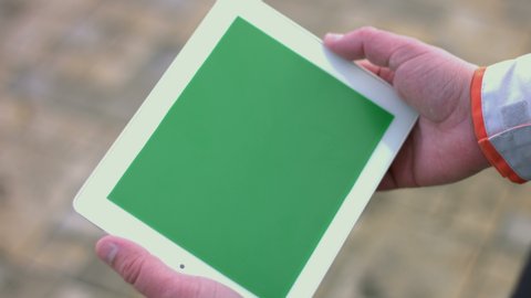 2 Shots: 1. Green screen of the tablet close-up. He presses and swipes the screen. 2. A man (worker, engineer) in overalls and protective helmet uses a tablet. Slow motion 2x.