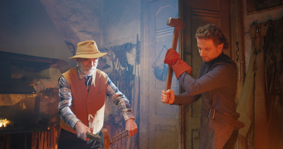 Two Caucasian blacksmiths, young and old men, working in antique forge and forging while beating with hammers on metal item. Producing of iron things. Smithy manufactory concept. Father and son work.