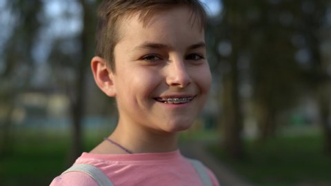 Portrait of cute boy looking camera in spring garden. Closeup smiling guy in brackets posing outdoors. Relaxed male teenager in teeth brackets staying outside. Young man smiling in park at sunset