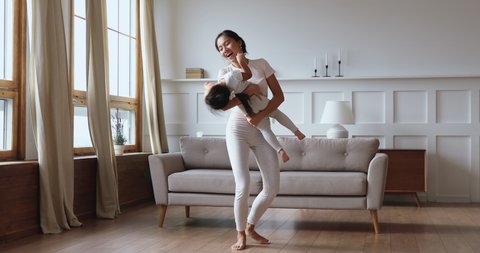 Emotional laughing young vietnamese mother lifting holding happy cute baby daughter, dancing barefoot in modern living room. Overjoyed mixed race asian family playing having fun together at home.