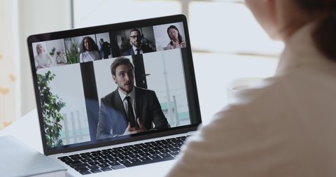 Male boss video conference calling corporate team people on laptop screen. Female distance employee talking with diverse colleagues group video conferencing in virtual webcam chat. Over shoulder view