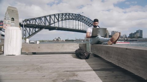 Man Using Laptop on Bench by Sydney Harbour Bridge. Low Angle Wide Shot