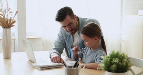 Dad helping cute small kid daughter doing homework at home. Young adult parent father or tutor explaining education teaching school girl pupil studying with daddy. Children remote education concept