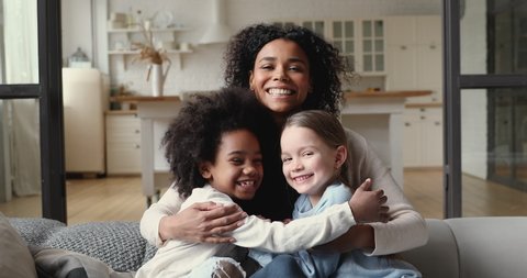 Happy affectionate mixed race family young adult mother hugging two cute diverse ethnicity children daughters and cuddling. Foster parent mum and small kids girls step sisters look at camera at home.