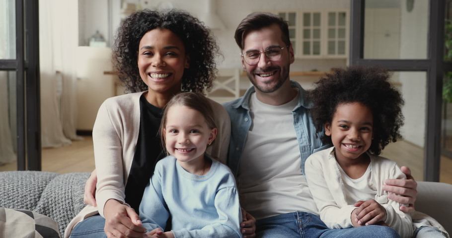 Cheerful interracial family young diverse ethnicity parents and kids looking at camera, laughing tickling on sofa at home. African mom, caucasian dad and mixed race children having fun in living room. | Shutterstock HD Video #1051507306