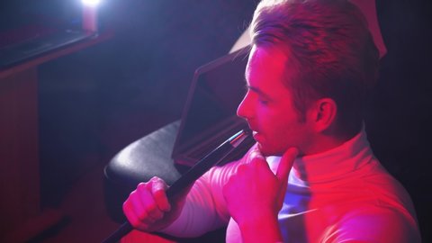 portrait of young man who smokes a hookah exhales smoke at a meeting with friends in a bar on a blue-red background side view