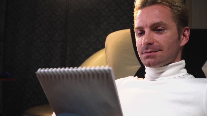 portrait of a man who writes his knowledge of thoughts in a notebook rejoices and smiles side view, sits on a light sofa
