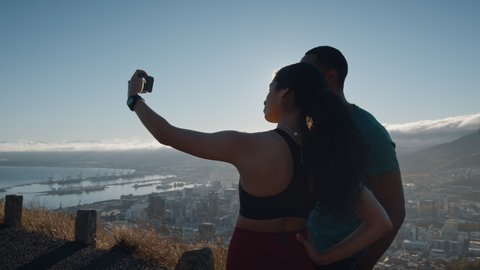 Woman taking a picture with man flexing biceps on a hillside road in the morning. Man and woman runner taking break after running workout on mountain road taking selfies with a mobile phone.
