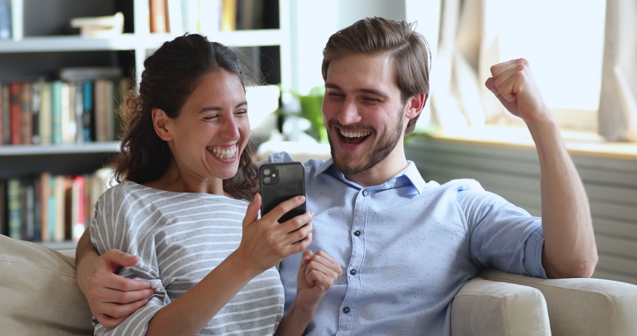 Overjoyed young couple looking at smart phone winning gift or prize in social media app sit on sofa at home. Excited happy millennial man and woman winners celebrating mobile victory together concept. Royalty-Free Stock Footage #1051518904