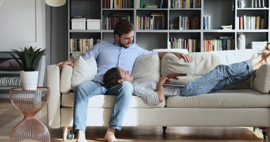 Relaxed millennial couple lounge on couch in living room using pc laptop together. Young smiling husband and wife resting on sofa enjoying browsing web on computer, talking and laughing at home. | Shutterstock HD Video #1051518910