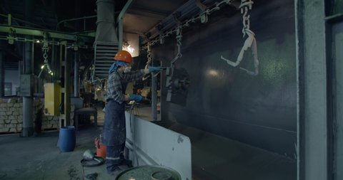 A girl in an orange helmet launches a metal part. At a machine-building plant