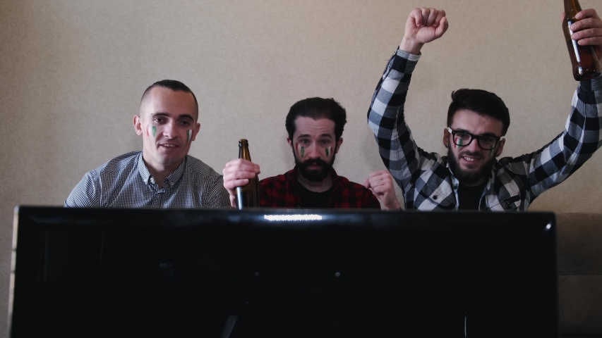 People are cheering for Italian team. Three men sit on sofa at home. Men raised hands up.