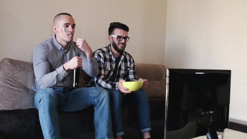 People are cheering for Italian team. Two men sit on sofa at home. Men drink beer and eat food during game.