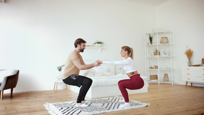 Young couple is doing squats excercises at home in cozy bright bedroom. Stay at home concept