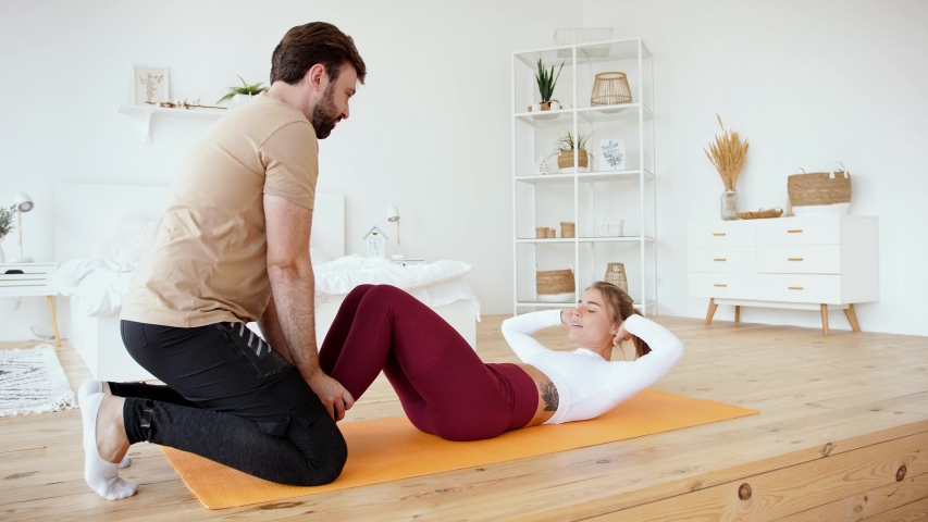 Young couple is doing sport excercises at home in cozy bright bedroom. Young man helps his girlfriend to do abs training. Stay at home concept