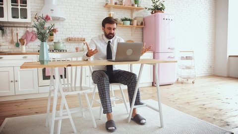 Young bearded man working from home with laptop wearing shirt, tie and pajama pants in kitchen
