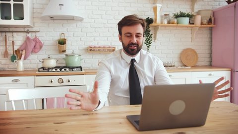 Young bearded man working from home with laptop and making a video call wearing shirt with tie in the kitchen, slow motion