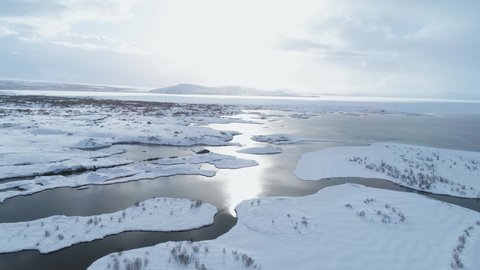 Aerial drone shot flying above Icelandic lake. Top view from sky over water, coast, white snow, mountain and sky with sun. Winter vacation in Thingvellir national park, Iceland, sunny day