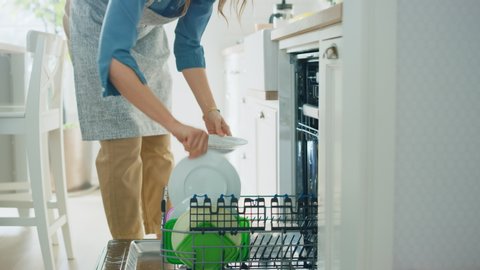 Beautiful Female is Loading Dirty Plates into a Dishwasher Machine in a Bright Sunny Kitchen. Girl in Wearing an Apron. Young Housewife Uses Modern Appliance to Keep the Home Clean.