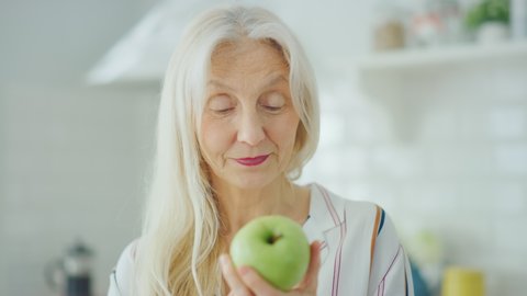 Authentic Portrait of Senior Female Pensioner in a Bright Kitchen at Home Eating an Apple. Beautiful Old Female with Gray Hair Poses for the Camera and Gently Smiles. Happy Vegetarian Full of Health.