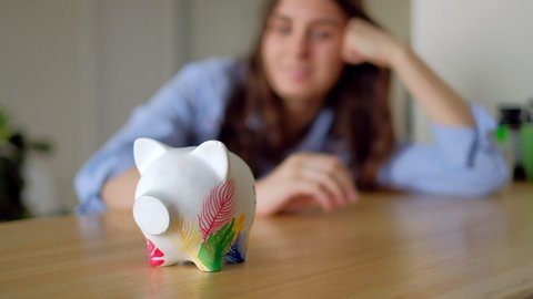 Young smiling woman sitting on a table and puts a coin in a piggy bank.