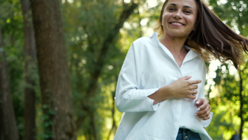 A beautiful smiling woman with green eyes bounces and poses for a good keepsake photograph in a city autumn park among the trees. Photo session in a warm sunny park for a pretty girl in a white shirt.